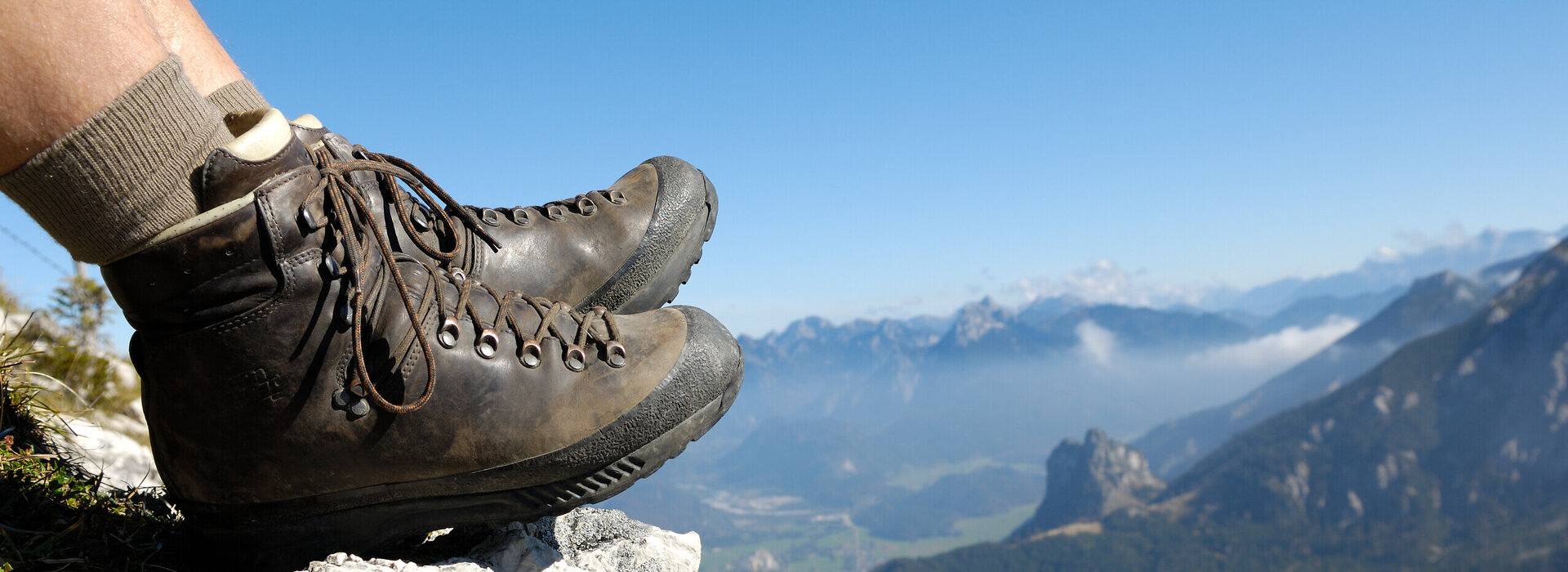 Das Bild zeigt eine Person, die auf einem Berg sitzt und nach unten blickt. Allerdings sind nur die ledernen Bergschuhe und die Wadern der Person zu sehen. Weiter unten erkennt man Bergwiesen, Wälder und ein Tal. Dahinter erheben sich weitere Berge. Der Himmel ist blau. Die Sonne scheint.  