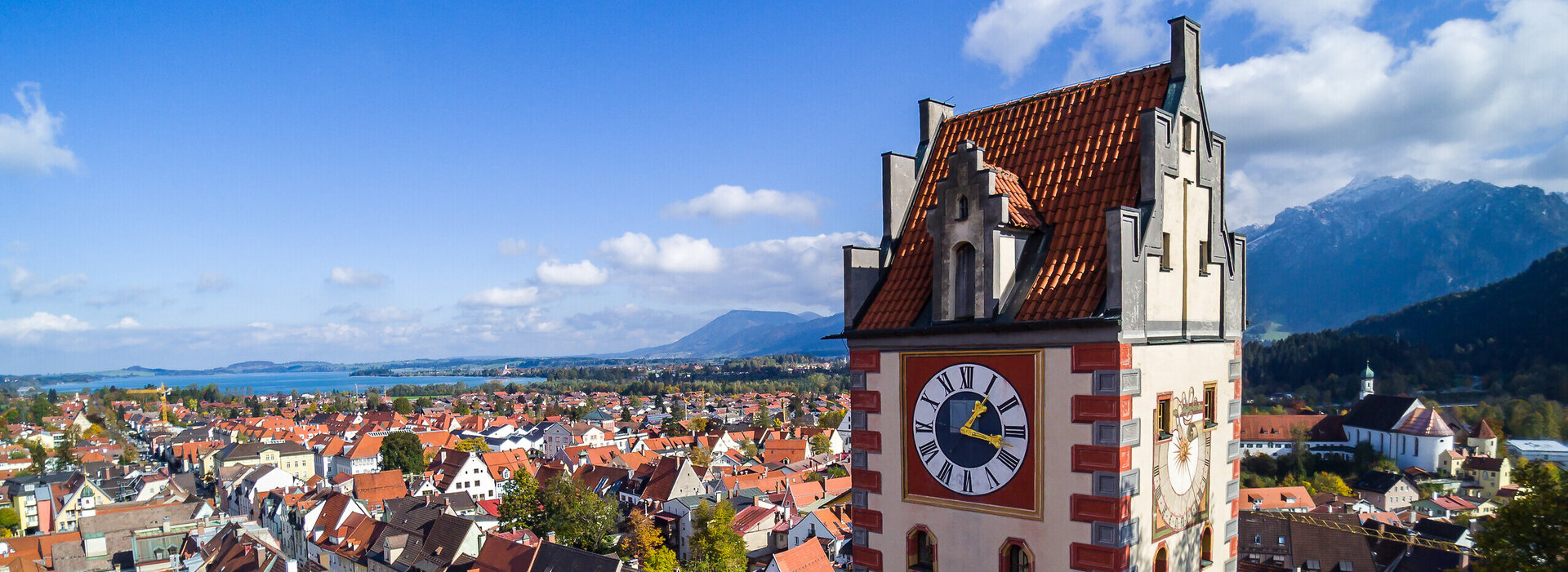 Das Foto zeigt Füssen von oben. Den Blick des Betrachters wird über die Dächer von Füssen geleitet. Im Vordergrund erhebt sich der sogenannte Uhrturm des Hohen Schlosses. Dahinter beginnen die Dächer der Füssener Altstadt. Im Hintergrund kann man schon die Berge und den türkisblauen Forggensee erkennen. 