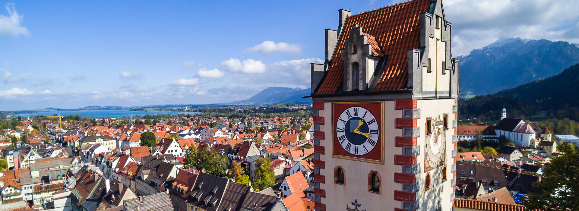 Das Foto zeigt Füssen von oben. Den Blick des Betrachters wird über die Dächer von Füssen geleitet. Im Vordergrund erhebt sich der sogenannte Uhrturm des Hohen Schlosses. Dahinter beginnen die Dächer der Füssener Altstadt. Im Hintergrund kann man schon die Berge und den türkisblauen Forggensee erkennen. 