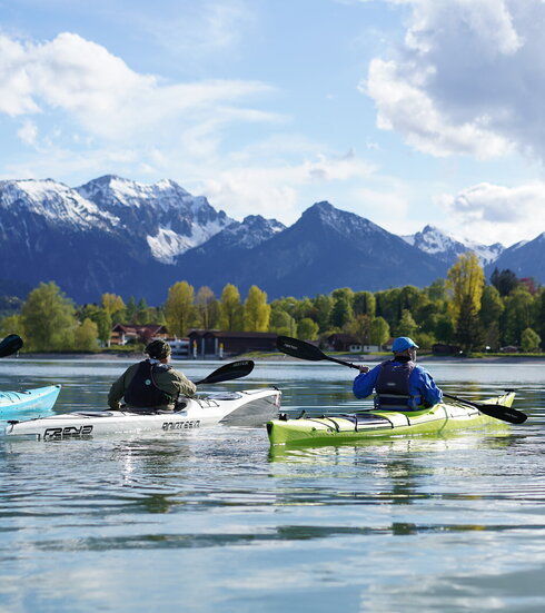 Das Bild zeigt drei Kajak-Fahrer, die auf einem See, nebeneinander in Richtung Ufer fahren. Sie haben dem Betrachter den Rücken zugewandt, tragen Sportkleidung und Sonnenbrillen. Am Ufer stehen hohe Bäume. Wenige Gebäude sind zu sehen. Im Hintergrund erheben sich schneebedeckte Berge. Der Himmel ist blau mit weißen Wolken durchzogen. Die Sonne scheint.  