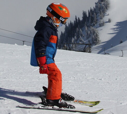 Das Bild fängt eine dynamische Wintersport-Szene ein. Im Zentrum des Bildes ist ein Skifahrer in Aktion zu sehen, der eine Spur durch eine schneebedeckte Berglandschaft zieht. Der Skifahrer trägt eine leuchtend blaue Jacke und orangefarbene Hosen, die sich vor dem weißen Schnee abheben. Der Sk Helm ist ein auffälliges Orange, das die Hosen ergänzt, und er trägt Schutzbrillen, um seine Augen vor der grellen Sonne zu schützen, die auf dem Schnee reflektiert.  Der Skifahrer ist nicht allein auf dem Berg. Im Hintergrund sind mehrere andere Skifahrer und Snowboarder zu sehen, die die Pisten genießen. Sie sind über die Piste verstreut, einige näher an der Kamera und andere weiter entfernt, was Tiefe in die Szene bringt.  Der Berg selbst ist dick mit Schnee bedeckt, mit der Skilift in der Ferne sichtbar. Der Himmel über ihm ist ein klarer Blau, der auf einen schönen Wintertag für Wintersportler hinweist. Die Gesamtszene ist eine der Aufregung und des Genusses an der Freiluft.