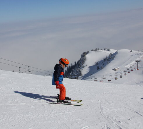 Das Bild fängt eine dynamische Wintersport-Szene ein. Im Zentrum des Bildes ist ein Skifahrer in Aktion zu sehen, der eine Spur durch eine schneebedeckte Berglandschaft zieht. Der Skifahrer trägt eine leuchtend blaue Jacke und orangefarbene Hosen, die sich vor dem weißen Schnee abheben. Der Sk Helm ist ein auffälliges Orange, das die Hosen ergänzt, und er trägt Schutzbrillen, um seine Augen vor der grellen Sonne zu schützen, die auf dem Schnee reflektiert.  Der Skifahrer ist nicht allein auf dem Berg. Im Hintergrund sind mehrere andere Skifahrer und Snowboarder zu sehen, die die Pisten genießen. Sie sind über die Piste verstreut, einige näher an der Kamera und andere weiter entfernt, was Tiefe in die Szene bringt.  Der Berg selbst ist dick mit Schnee bedeckt, mit der Skilift in der Ferne sichtbar. Der Himmel über ihm ist ein klarer Blau, der auf einen schönen Wintertag für Wintersportler hinweist. Die Gesamtszene ist eine der Aufregung und des Genusses an der Freiluft.