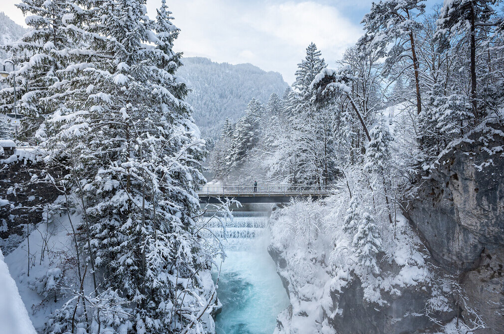 Das Bild zeigt einen eisblauen Fluss, der sich seinen Weg durch eine Felsschlucht bahnt. Über den Fluss führt eine Brücke. Die Felsen und die weitere Umgebung sind verschneit. Der Himmel ist blau.   