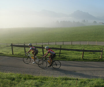 Das Bild zeigt zwei Rennradfahrer in Trikots und Radlerhosen, die einen Hügel emporfahren. Beide tragen einen Helm. Sie sind gerade um eine enge Kurve gefahren. Die Straße ist geteert. An die Straße grenzen Wiesen an. Parallel zur Straße verläuft ein Holzzaun. Der Morgennebel lichtet sich langsam. Die Sonne scheint. Der Himmel ist blau. Im Hintergrund erheben sich die Berge.  