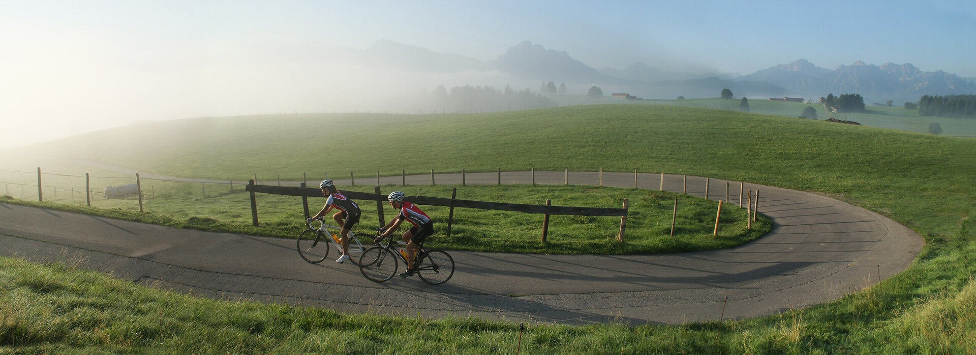 Das Bild zeigt zwei Rennradfahrer in Trikots und Radlerhosen, die einen Hügel emporfahren. Beide tragen einen Helm. Sie sind gerade um eine enge Kurve gefahren. Die Straße ist geteert. An die Straße grenzen Wiesen an. Parallel zur Straße verläuft ein Holzzaun. Der Morgennebel lichtet sich langsam. Die Sonne scheint. Der Himmel ist blau. Im Hintergrund erheben sich die Berge.  