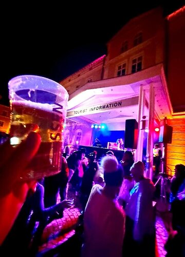 Das Bild zeigt eine Hausfassade. Unter dem überdachten Haupteingang steht ein beleuchtets DJ-Pult. Darüber hängt ein Schild Tourist Information. Viele Leute tanzen. Sie tragen Mützen und Winterkleidung. Im Vordergrund hält eine Hand einen Becher mit Bier gefüllt. Auf dem Becher steht „Füssen“.  