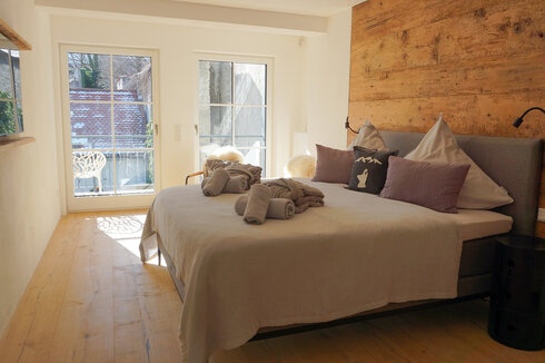 Das Bild zeigt ein lichtdurchflutetes Hotelzimmer, dass mit einem Doppelbett und zwei Nachtkästchen möbliert ist. Die Wand, an dem das Bett steht ist aus Holz. Über dem Bett liegen eine Tagesdecke, zusammengerollte Handtücher und zwei zusammengelegte Morgenmäntel. Im Hintergrund ist durch die Fenster ein Balkon zu erkennen auf dem ein Stuhl steht. 