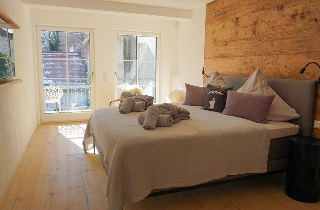 Das Bild zeigt ein lichtdurchflutetes Hotelzimmer, dass mit einem Doppelbett und zwei Nachtkästchen möbliert ist. Die Wand, an dem das Bett steht ist aus Holz. Über dem Bett liegen eine Tagesdecke, zusammengerollte Handtücher und zwei zusammengelegte Morgenmäntel. Im Hintergrund ist durch die Fenster ein Balkon zu erkennen auf dem ein Stuhl steht. 