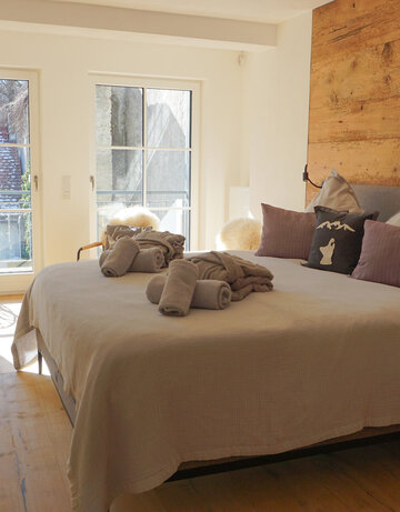 Das Bild zeigt ein lichtdurchflutetes Hotelzimmer, dass mit einem Doppelbett und zwei Nachtkästchen möbliert ist. Die Wand, an dem das Bett steht ist aus Holz. Über dem Bett liegen eine Tagesdecke, zusammengerollte Handtücher und zwei zusammengelegte Morgenmäntel. Im Hintergrund ist durch die Fenster ein Balkon zu erkennen auf dem ein Stuhl steht. 