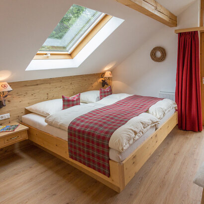 Das Bild zeigt ein lichtdurchflutetes Hotelzimmer. Das Doppelbett ist weiß überzogen. Auf den Bettdecken liegt rot und grau karierte Tagesdecke. Die Wand, an der das Bett steht, ist aus Holz. Daran hängen zwei Nachtischlampen in Form von Hirschköpfen. Auch die gegenüberliegende Wand ist aus Holz. Davor steht ein Schreibtisch. Darüber hängt ein Fernseher. Durch die geschlossene Balkontür blickt man auf grüne Bäume. Das Zimmer hat eine Dachschräge über dem Bett. Das Dachfenster direkt über dem Doppelbett.  