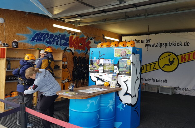Das Bild zeigt eine lebhafte Szene in einem Sportgeschäft. Das Geschäft ist vollgepackt mit verschiedenen Sportausrüstungen, einschließlich eines großen blauen und weißen Kühlschranks, eines roten Feuerlöschers und eines gelben Helms. Das Geschäft ist geschäftig, mit mehreren Menschen, die die verschiedenen Artikel durchstöbern. Die Wände des Geschäfts sind mit bunten Graffiti verziert, was der Atmosphäre eine lebendige Note verleiht. Der Name des Geschäfts "ALPITZ KICK" ist prominent angebracht, was darauf hinweist, dass sich das Geschäft auf Fußballausrüstung spezialisiert hat. Die Adresse des Geschäfts, "www.alpitzkick.de", deutet darauf hin, dass es sich auch um einen Online-Store handelt. Insgesamt suggeriert die Szene ein gut sortiertes und sehr beliebtes Sportgeschäft, das Kunden mit seiner umfangreichen Auswahl an Sportartikeln und dem einzigartigen Flair anlockt.