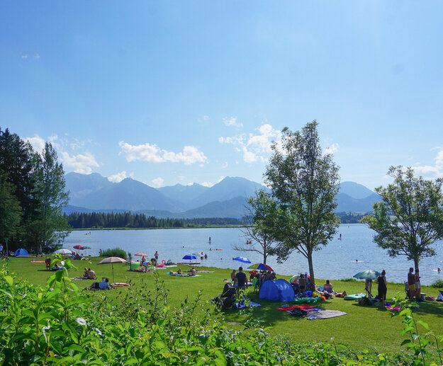 Das Bild zeigt einen See. Eine grüne Wiese grenzt an den See. Auf der Wiese liegen zahlreiche Personen auf Decken und Handtüchern unter bunten Sonnenschirmen. Im Wasser schwimmen Menschen oder paddelt auf SUPs. Im Hintergrund erheben sich die Berge. Der Himmel ist blau. Die Sonne scheint. 