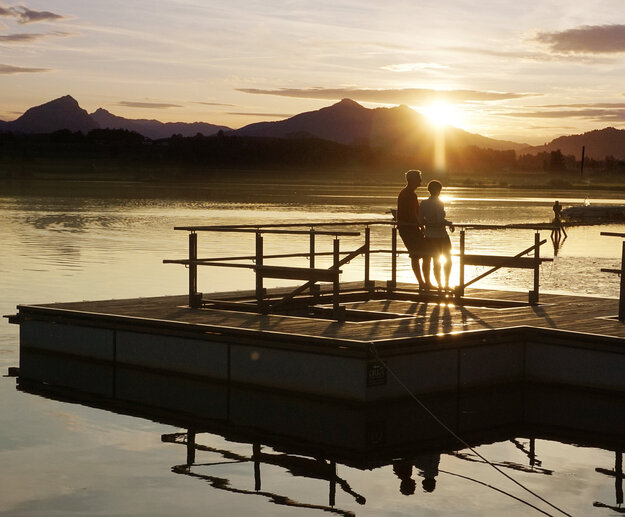 Das Bild zeigt zwei Personen, die auf einer hölzernen Plattform stehen, die auf einem See schwimmt. Die untergehende Sonne taucht die Szenerie in ein warmes Licht. Im Hintergrund sind die Silhouetten hoher Berge zu sehen.
