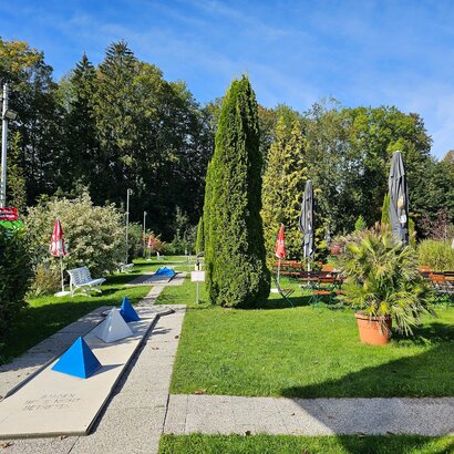 Auf dem Bild sind mehrere Minigolfbahnen zu sehen. Es gibt eine lockere Bepflanzung Sitzgelegenheiten und rote Sonnenschirme. Das Wetter ist sonnig und wolkenlos.