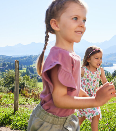Das Bild fängt einen ruhigen Moment in einem Park ein. Die beiden jungen Mädchen sind der Hauptfokus des Bildes. Das Mädchen in der Vordergrund trägt ein pinkes Oberteil und hat ihre Haare zu einem Zopf zusammengebunden. Sie geht auf die Kamera zu, ihr Gesicht erstrahlt von einem Lächeln. Das andere Mädchen, das ein weißes Kleid mit pinken Blumen trägt, steht ein wenig entfernt. Sie lächelt ebenfalls, ihr Blick ist auf die Kamera gerichtet.  Der Park, in dem sie sich befinden, ist üppig grün mit einem Pfad, der zu einem Holzzaun führt. Jenseits des Zauns ist ein schöner See zu sehen, dessen ruhiges Wasser den klaren blauen Himmel darüber widerspiegelt. Die Berge in der Ferne verleihen der Szene eine friedvolle Ruhe. Die Freude der Mädchen und die friedliche Umgebung erzeugen ein angenehmes Zusammenspiel von jugendlicher Energie und natürlicher Schönheit.
