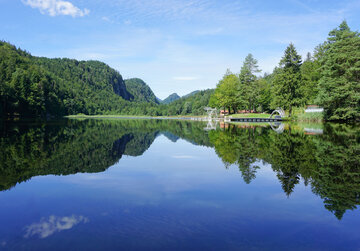 Das Bild zeigt eine ruhige Seenlandschaft. Der Obersee, der der zentrale Fokus des Bildes ist, ist von üppigem Grün umgeben und von Bergen gesäumt. Die ruhige Wasseroberfläche des Sees spiegelt die Umgebung wider. Auf der rechten Seite des Bildes befindet sich ein Steg mit einem Sprungturm. Der Himmel darüber ist klar und blau.