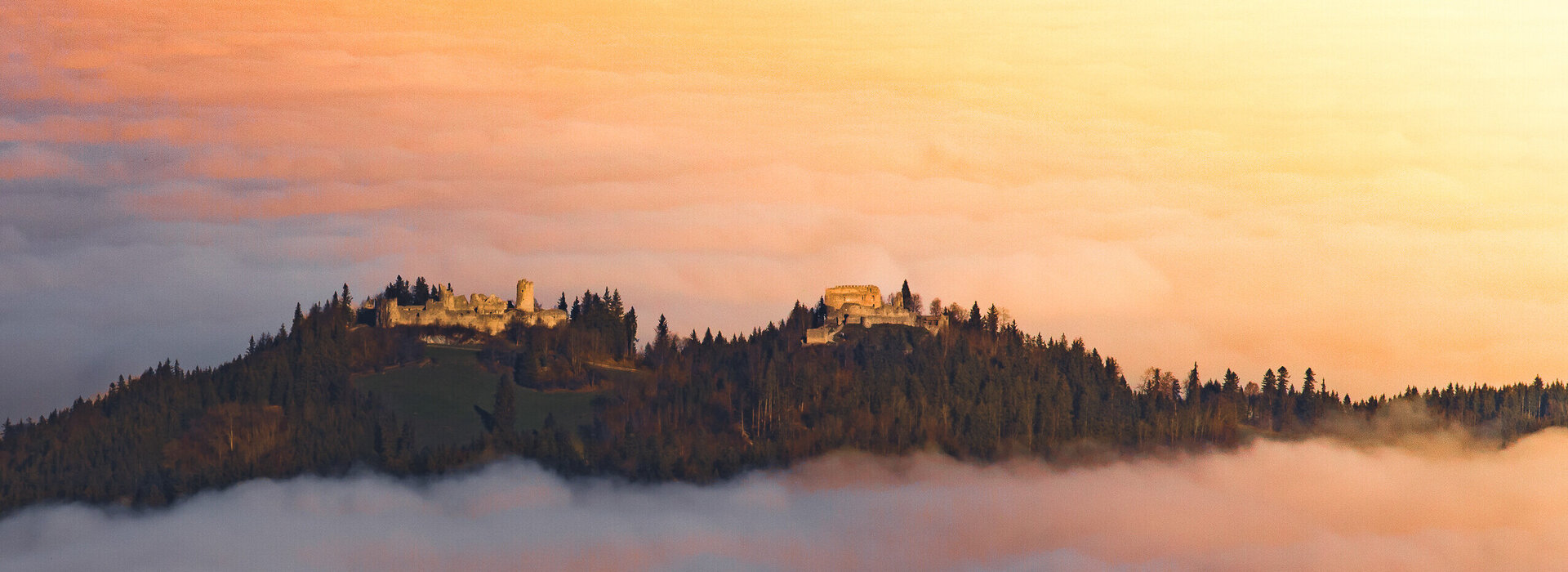 Das Bild fängt eine atemberaubende Aussicht einer Burg ein, die auf einem Hügel thront und von einem üppigen Wald umgeben ist. Die Burg, mit ihrer majestätischen Architektur, hebt sich vor dem Hintergrund des Himmels ab. Der Himmel ist in Tönen von Orange und Rosa gemalt, was darauf hindeutet, dass die Sonne gerade untergeht und die ganze Szene in ein warmes Licht taucht. Der auf dem Hügel liegende Burgberg ist mit Nebel bedeckt, was der Szene etwas Mystisches verleiht.