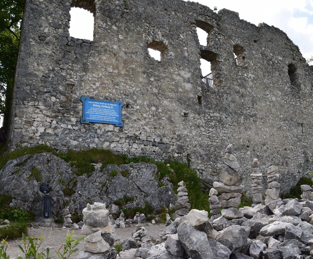 Das Bild zeigt eine majestätische Burg ähnliche Struktur, erbaut aus grauem Stein. Die Burg zeichnet sich durch ihre vielen Fenster und ein großes, blaues Schild aus, das auffällig an der Seitenfront des Gebäudes angebracht ist. Die Burg liegt auf einem Hügel, umgeben von einer üppigen grünen Landschaft. Der Himmel über ihr ist ein klarer blauer Himmel, der die ganze Szene ästhetisch ansprechend gestaltet. Die Größe und Schönheit der Burg und die friedliche Umgebung machen einen faszinierenden Anblick aus.