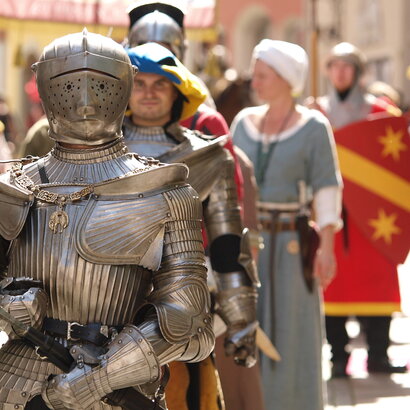 Das Bild erfasst eine lebhafte Szene eines mittelalterlich-thematisierten Festes. Das Hauptaugenmerk liegt auf einem Mann in einer vollständigen Rüstung, inklusive Helm und Schwert. Er steht in der Mitte des Bildes und zieht die Aufmerksamkeit des Betrachters auf sich. Zu seiner Linken befindet sich eine Frau in mittelalterlichem Gewand und hält einen Schild. Zu seiner Rechten steht ein weiterer Mann in Rüstung, der ein Banner hält. Im Hintergrund sind andere Personen in mittelalterlichem Gewand zu sehen, die zur festlichen Stimmung beitragen. Die Gebäude im Hintergrund sowie die Kostüme zeigen die mittelalterlichen Häuser der Altstadt von Füssen. Die gesamte Szene ist ein lebhafter Ausdruck mittelalterlicher Kultur und Geschichte.