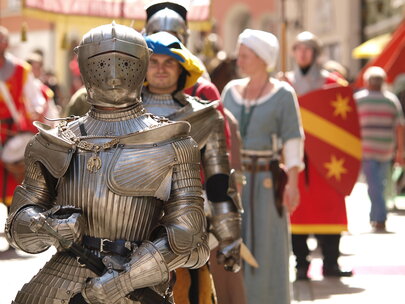 Das Bild erfasst eine lebhafte Szene eines mittelalterlich-thematisierten Festes. Das Hauptaugenmerk liegt auf einem Mann in einer vollständigen Rüstung, inklusive Helm und Schwert. Er steht in der Mitte des Bildes und zieht die Aufmerksamkeit des Betrachters auf sich. Zu seiner Linken befindet sich eine Frau in mittelalterlichem Gewand und hält einen Schild. Zu seiner Rechten steht ein weiterer Mann in Rüstung, der ein Banner hält. Im Hintergrund sind andere Personen in mittelalterlichem Gewand zu sehen, die zur festlichen Stimmung beitragen. Die Gebäude im Hintergrund sowie die Kostüme zeigen die mittelalterlichen Häuser der Altstadt von Füssen. Die gesamte Szene ist ein lebhafter Ausdruck mittelalterlicher Kultur und Geschichte.