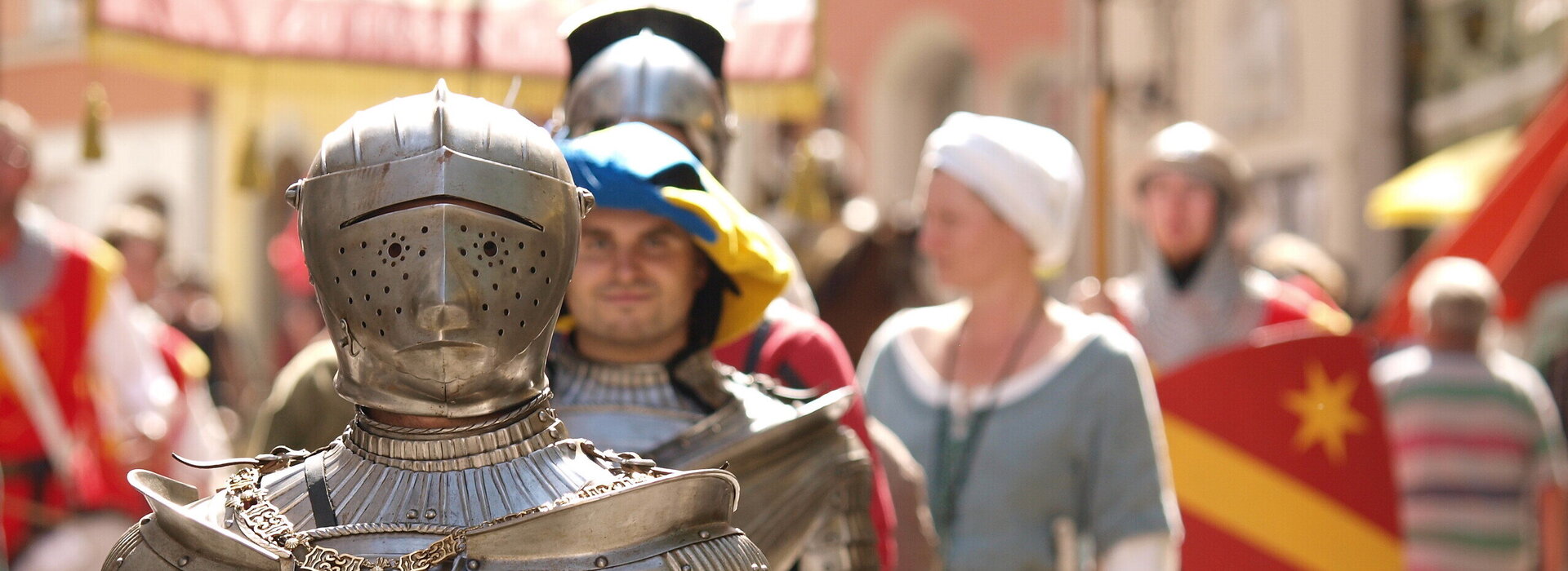 Das Bild erfasst eine lebhafte Szene eines mittelalterlich-thematisierten Festes. Das Hauptaugenmerk liegt auf einem Mann in einer vollständigen Rüstung, inklusive Helm und Schwert. Er steht in der Mitte des Bildes und zieht die Aufmerksamkeit des Betrachters auf sich. Zu seiner Linken befindet sich eine Frau in mittelalterlichem Gewand und hält einen Schild. Zu seiner Rechten steht ein weiterer Mann in Rüstung, der ein Banner hält. Im Hintergrund sind andere Personen in mittelalterlichem Gewand zu sehen, die zur festlichen Stimmung beitragen. Die Gebäude im Hintergrund sowie die Kostüme zeigen die mittelalterlichen Häuser der Altstadt von Füssen. Die gesamte Szene ist ein lebhafter Ausdruck mittelalterlicher Kultur und Geschichte.