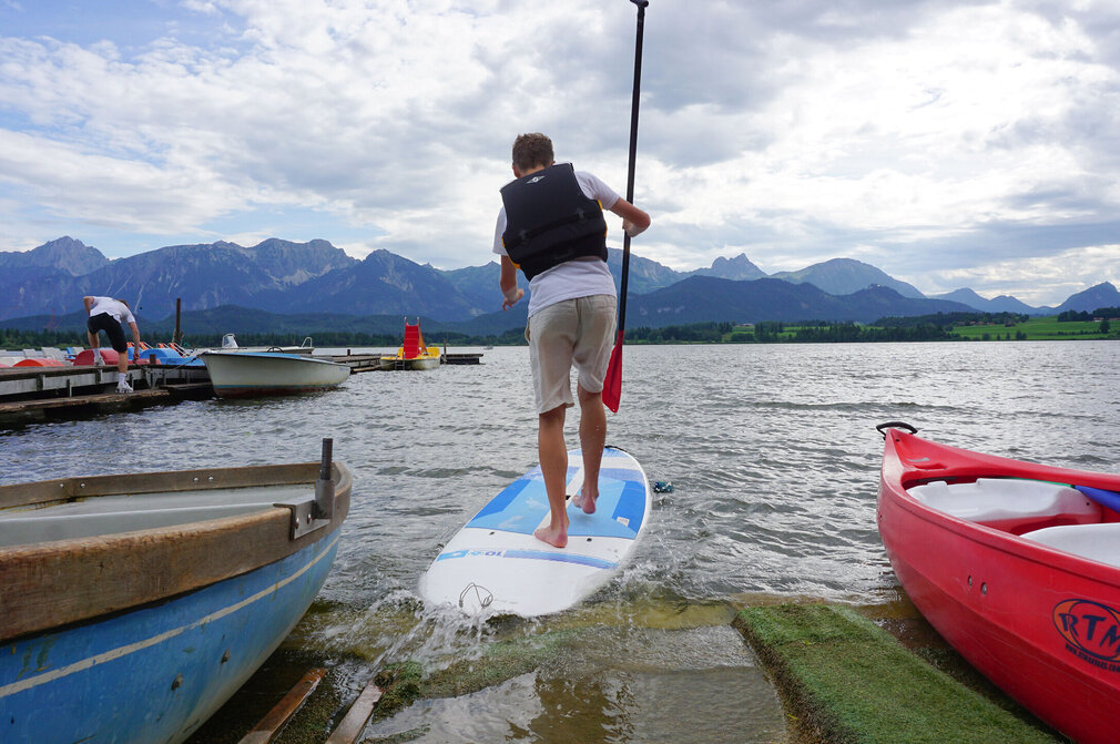 Das Bild fängt eine Szene an einer Anlegestelle ein. Ein Mann steht auf einem weißen Surfbrett, das auf einer Rampe positioniert ist. Er hält ein Paddel. Die Anlegestelle ist von mehreren Booten umgeben. Die Kulisse des Bildes ist ein schöner Bergkamm, der zur Ruhe der Szene beiträgt. Der Mann scheint sich darauf vorzubereiten, zu surfen, da er auf dem Surfbrett steht und in Richtung des Wassers blickt. Die Gesamtatmosphäre des Bildes ist friedlich und einladend, mit dem Mann, dem Surfbrett und den Booten, die alle zu einem Gefühl von Abenteuer und Entspannung beitragen.