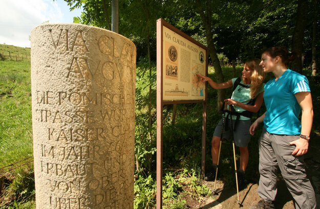 Das Bild zeigt zwei Frauen, die vor einer Tafel stehen. Auf der Tafel ist „Via Claudia Augusta“ zu lesen sowie ein Erklärungstext und unterschiedliche Abbildungen. Daneben steht eine Steinsäule auf der „Via Claudia Augusta“ unter weitere leider aus dieser Perspektive unleserliche Texte stehen. Dahinter erhebt sich ein grüner Hügel. Hinter den Frauen wachsen hohe Bäume. Die Sonne scheint.