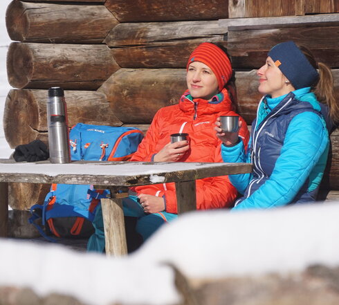Das Bild zeigt zwei Frauen, die an einem Holztisch vor einem Holzstadel sitzen. Nur ein Detail der Wand ist zu sehen. Beide tragen Winterkleidung und Stirnbänder. Sie haben je einen Thermoskannenbecher in der Hand. Die Thermoskannen stehen auf dem Tisch. Die Sonne scheint. Ringsum ist alles schneebedeckt. 