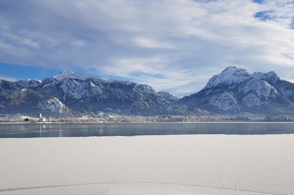 Das Bild zeigt ein atemberaubendes Panorama eines schneebedeckten Gebirgszuges unter einem klaren blauen Himmel. Die Berge, mit ihren zackigen Gipfeln, erheben sich majestätisch im Hintergrund. Die Vordergrunddomäne wird von einem stillen See dominiert, dessen ruhiges Wasser die Berge widerspiegelt. Die Oberfläche des Sees ist unberührt, was zur Ruhe des Bildes beiträgt. Die Gesamtkomposition des Bildes suggeriert eine friedliche und ruhige Landschaft, unberührt von menschlichem Eingriff. 