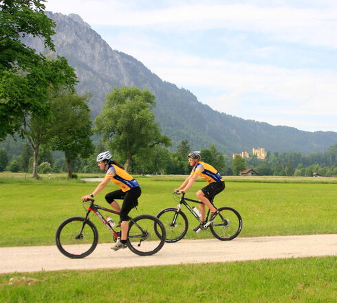 Das Bild zeigt einen Mann und eine Frau beim Fahrradfahren. Sie fahren auf Mountainbikes, tragen gelbe Trikots, schwarze Radlerhosen und Helme. Sie fahren in Richtung des linken Bildrands auf einem unbefestigten Feldweg, der von Wiesen umgeben ist. Im Hintergrund erheben sich schneebedeckte Berge und grün, bewaldete Hügel. Auf einem der Hügel thront das gelbe Schloss Hohenschwangau. Der Himmel ist blau. Die Sonne scheint. 