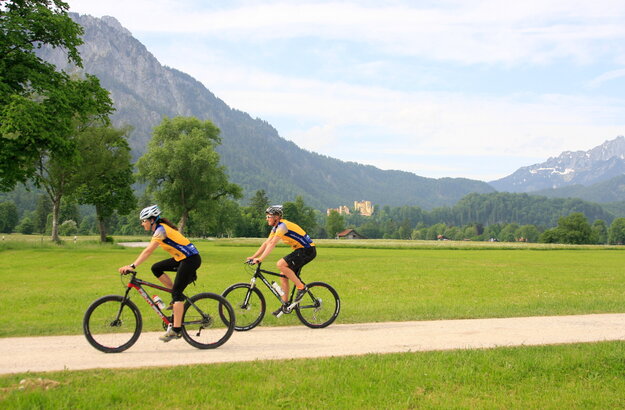 Das Bild zeigt einen Mann und eine Frau beim Fahrradfahren. Sie fahren auf Mountainbikes, tragen gelbe Trikots, schwarze Radlerhosen und Helme. Sie fahren in Richtung des linken Bildrands auf einem unbefestigten Feldweg, der von Wiesen umgeben ist. Im Hintergrund erheben sich schneebedeckte Berge und grün, bewaldete Hügel. Auf einem der Hügel thront das gelbe Schloss Hohenschwangau. Der Himmel ist blau. Die Sonne scheint. 
