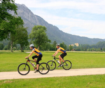 Das Bild zeigt einen Mann und eine Frau beim Fahrradfahren. Sie fahren auf Mountainbikes, tragen gelbe Trikots, schwarze Radlerhosen und Helme. Sie fahren in Richtung des linken Bildrands auf einem unbefestigten Feldweg, der von Wiesen umgeben ist. Im Hintergrund erheben sich schneebedeckte Berge und grün, bewaldete Hügel. Auf einem der Hügel thront das gelbe Schloss Hohenschwangau. Der Himmel ist blau. Die Sonne scheint. 