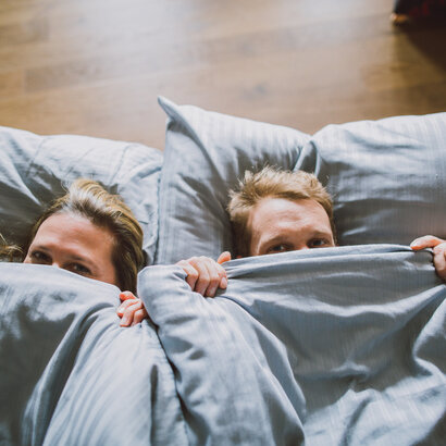Das Bild zeigt eine Frau und einen Mann von oben, die sich eine blaugraue Bettdecke über die Nasenspitze ziehen und in die Kamera blicken. Ihre Köpfe sind auf ebensoblaugrauen Kissen gebettet.