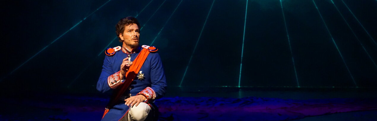 Das Foto zeigt eine Mann mit einer historischen blauen Uniform, weißen Hosen und roten Reitstiefeln. Er trägt zudem eine rote Schärpe und einen Orden. Der Mann kniet auf dem Boden. Es handelt sich um König Ludwig II. von Bayern in einer Szene aus dem Musical Ludwig² - Der König ist zurück. 