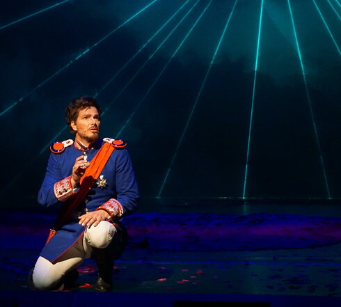 Das Foto zeigt eine Mann mit einer historischen blauen Uniform, weißen Hosen und roten Reitstiefeln. Er trägt zudem eine rote Schärpe und einen Orden. Der Mann kniet auf dem Boden. Es handelt sich um König Ludwig II. von Bayern in einer Szene aus dem Musical Ludwig² - Der König ist zurück. 