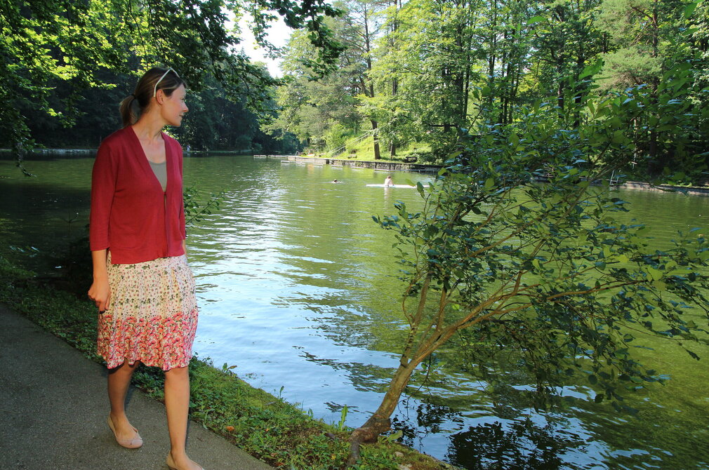 Das Bild zeigt eine Frau auf einem asphaltierten Weg am Ufer eines grünen Sees. Sie ist in einem lebhaften roten Pullover und einem floralen Rock gekleidet. Ihr Blick ist auf den See gerichtet, wo mehrere Personen schwimmen. Der See ist von üppigen grünen Bäumen umgeben und schafft eine ruhige Atmosphäre. 