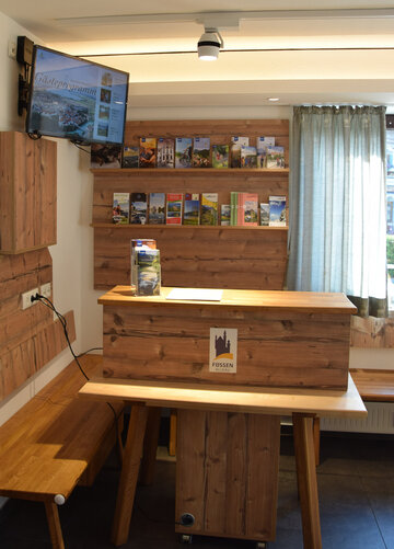 Das Bild zeigt ein Detail eines Raumes. Vor einer Holzeckbank steht ein hölzerner Tresen. An der Wand dahinter ist ein Prospektregal angebracht, das mit zahleichen Prospekten gefüllt ist. An der Nebenwand des Tresens ist ein Monitor angebracht. 