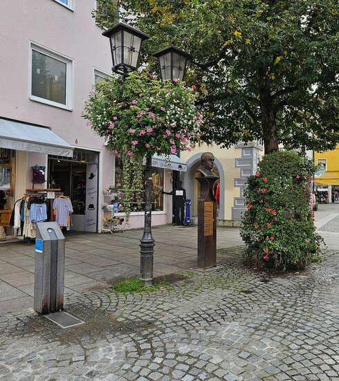 Das Bild zeigt einen Trinkbrunnen in der Fußgängerzone von Füssen. Im Hintergrund sind die bunten Fassaden der Stadt zu sehen. Neben dem Trinkbrunnen steht eine bronzene Säule, die die Büste von Sebastian Kneipp trägt. Im Hintergrund sind viele Schaufenster zu sehen. Die Sonne scheint.  