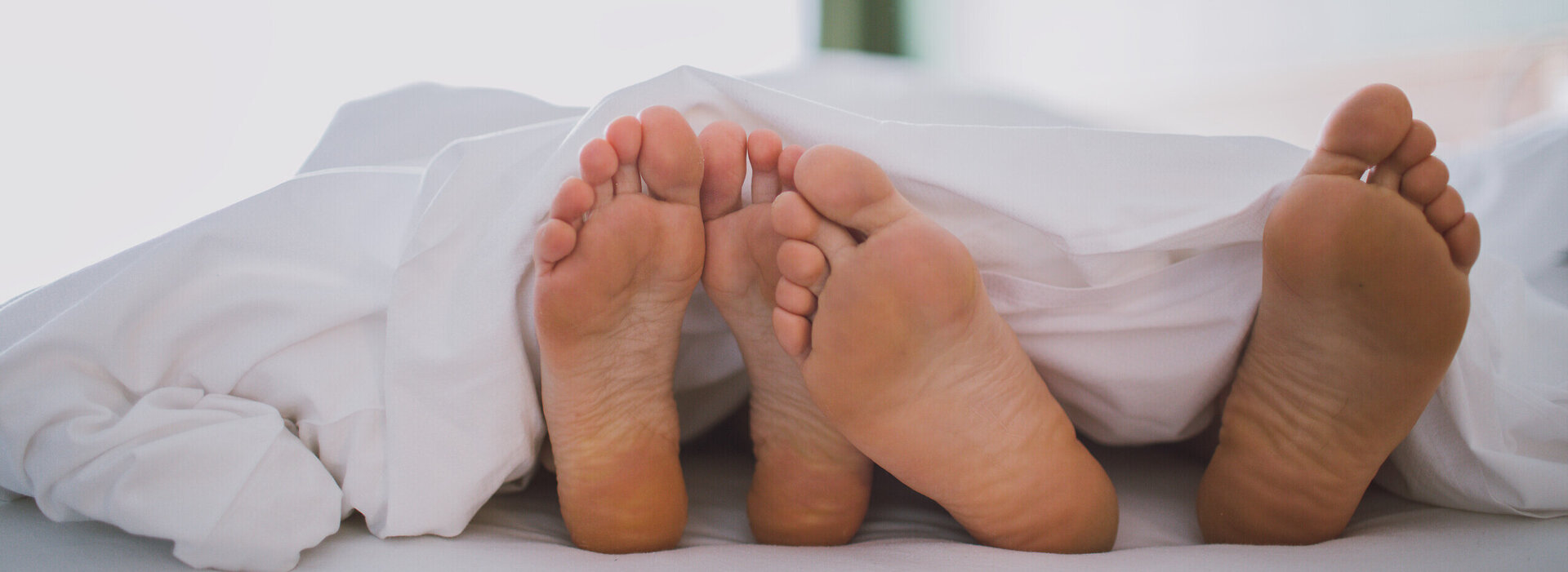 Das Bild zeigt zwei nackte Fußpaare, die unter einer weißen Decke hervorschauen. Die Füße sind so positioniert, dass sie fast verborgen sind. Die Decke, die das Hauptmotiv des Bildes ist, ist ordentlich verstaut, was eine behagliche und einladende Atmosphäre schafft. Die Kulisse ist verschwommen, was die Hauptmotive - die Füße und die Decke - betont. Die Gesamtzusammenstellung des Bildes suggeriert einen Moment der Entspannung und des Wohlbehagens