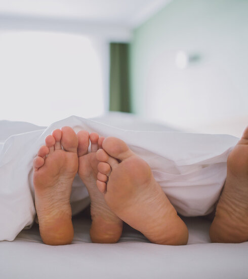 Das Bild zeigt zwei nackte Fußpaare, die unter einer weißen Decke hervorschauen. Die Füße sind so positioniert, dass sie fast verborgen sind. Die Decke, die das Hauptmotiv des Bildes ist, ist ordentlich verstaut, was eine behagliche und einladende Atmosphäre schafft. Die Kulisse ist verschwommen, was die Hauptmotive - die Füße und die Decke - betont. Die Gesamtzusammenstellung des Bildes suggeriert einen Moment der Entspannung und des Wohlbehagens