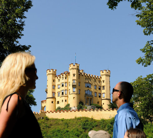 Das Bild zeigt eine friedliche Szene einer Familie bestehend aus einer Frau, einem Mann und zwei Kindern, die vor einem Schloss stehen. Die Frau, in einem schwarzen Oberteil gekleidet, steht links im Bild. Sie schaut in Richtung rechts, ihr Blick folgt dem Mann und den zwei Kindern. Der Mann trägt ein blaues Hemd und steht in der Mitte des Bildes, flankiert von den beiden Kindern, das eine in einem orangefarbenen Hemd und das andere in einem blauen Hemd. Das Schloss, das farblich in einem leuchtenden Gelb gestaltet ist, steht majestätisch im Hintergrund. Die Familie scheint einen Ausflug zu genießen und die Ansichten des Schlosses zu bewundern.