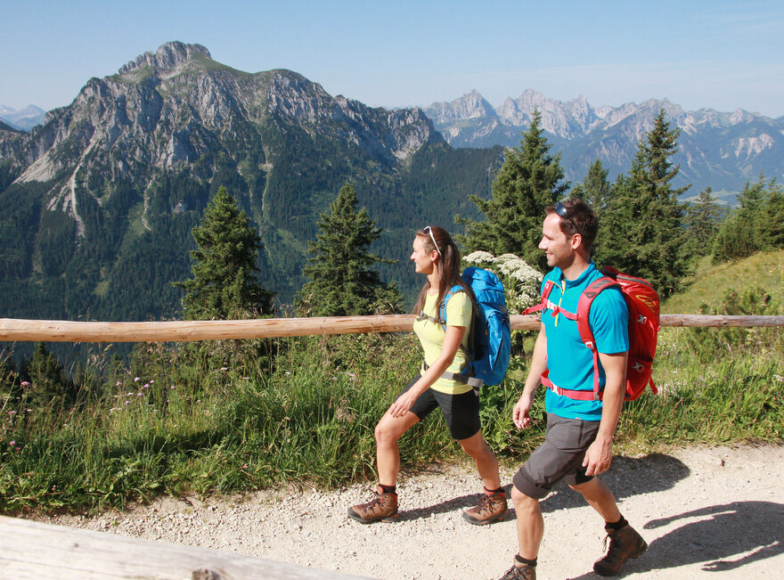 Das Bild zeigt einen Mann und eine Frau in kurzer Wanderkleidung in auf einem unbefestigten Weg laufen. Parallel zum Kiesweg verläuft ein Holzgeländern. Im Hintergrund erheben sich die Berge. Der Himmel ist blau. Die Sonne scheint. 