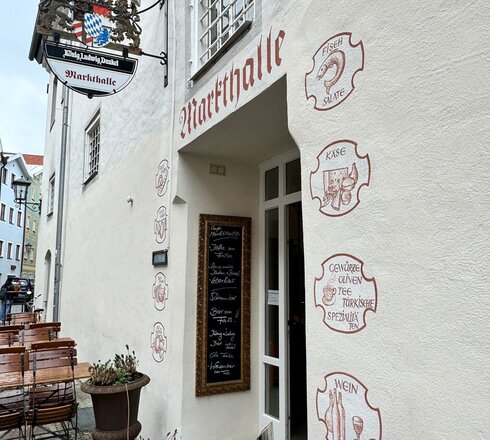 Das Bild zeigt eine verzauberte europäische Straßenszene. Der Hauptaugenmerk liegt auf einem weißem Gebäude mit einem roten Vordach. Das Gebäude ist mit einem Schild geschmückt, das in roten Buchstaben den Namen "Wirtshalle" trägt. Die Fassade des Gebäudes ist mit verschiedenen Zeichen und Symbolen verziert, darunter ein Fisch, ein Weinglas und eine Kaffeetasse. Diese Zeichen lassen darauf schließen, dass das Gebäude ein Restaurant oder Café beherbergt.  Vor dem Gebäude stehen mehrere Holztische und -stühle, die Vorbeigehende einladen, sich hinzusetzen und die Atmosphäre zu genießen. Die Straße selbst ist von anderen Gebäuden umgeben, die zur märchenhaften europäischen Stimmung beitragen. Die Gesamtszene ist eine friedvolle, genußorientierte Tag in einem europäischen Städtchen.