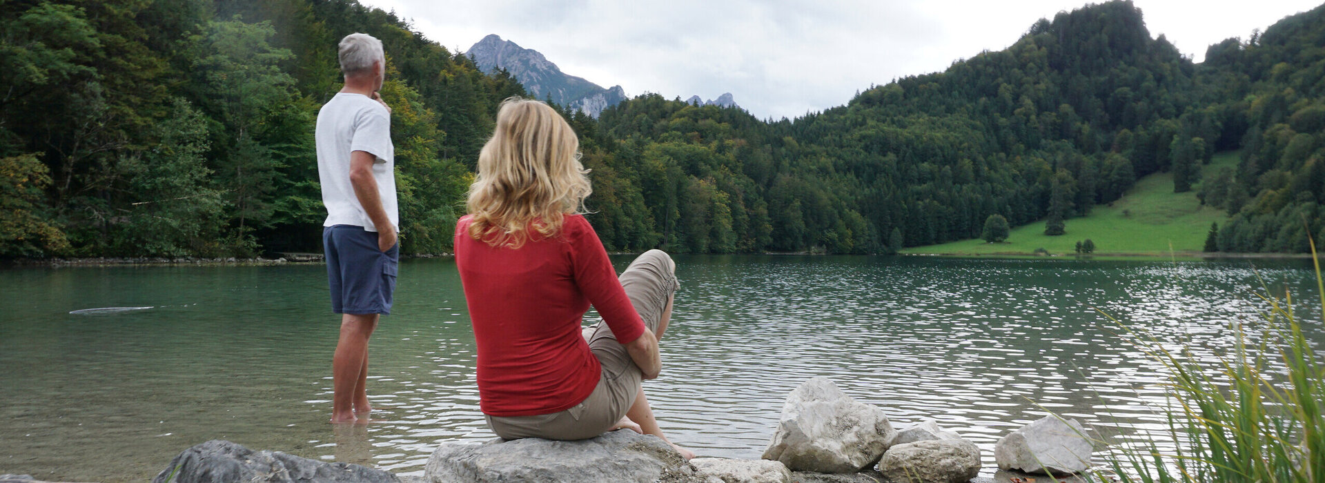 Man blickt auf eine Frau und einen Mann die auf einen See blicken. Die Frau sitzt auf einem großen Stein am Ufer. Der Mann steht im köcheltiefen, ufernahen Wasser. Der See ist ringsum mit grünen Wäldern umgeben. Dahinter erheben sich die Berge. Der Himmel ist wolkenverhangen. 