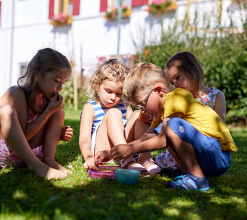 Das Bild zeigt eine herrliche Szene von vier Kindern, die in einer wunderschönen Wiese spielen. Die Kinder, zwei Mädchen und zwei Jungen, sind alle elegant in Sommerkleidung gekleidet. Das Mädchen auf der linken Seite sitzt mit überschlagenen Beinen, ihre Aufmerksamkeit ist auf die anderen drei Kinder gerichtet. Das Mädchen in der Mitte sitzt auf der Wiese, ihr Blick ist auf den Jungen in der Mitte gerichtet. Der Junge in der Mitte kniet auf der Wiese, seine Hände sind mit einer Aufgabe beschäftigt. Der Junge auf der rechten Seite sitz auf der Wiese und richtet seine Aufmerksamkeit auf das Mädchen in der Mitte. Die Wiese, auf der sie spielen, ist ein lebhaftes Grün, durchsetzt mit ein paar Kübelpflanzen. Im Hintergrund ist ein weißes Gebäude mit roten Fensterläden zu sehen, das eine Farbtupfer in die Szene bringt. Die Freude der Kinder und die Ruhe der Umgebung ergeben das Bild eines perfekten Sommertages.