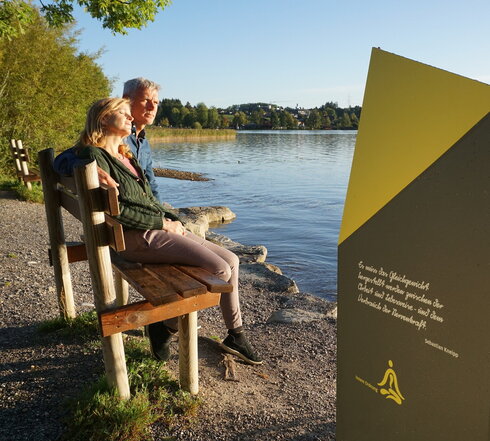 Das Bild zeigt einen Mann und eine Frau, die auf einer Holzbank sitzen. Die Frau lehnt sich an die Schulter des Mannes und hat die Augen geschlossen. Die Bank steht am Ufer eines Sees. Die Abendsonne taucht die Szenerie in warmes Licht. Im Vordergrund steht eine grau gelbe Metallsäule auf der zu lesen ist: „Es muss das Gleichgewicht hergestellt werden zwischen der Arbeit und Lebensweise – und dem Verbrauch der Nervenkraft. – Sebastian Kneipp, Innere Ordnung“. 