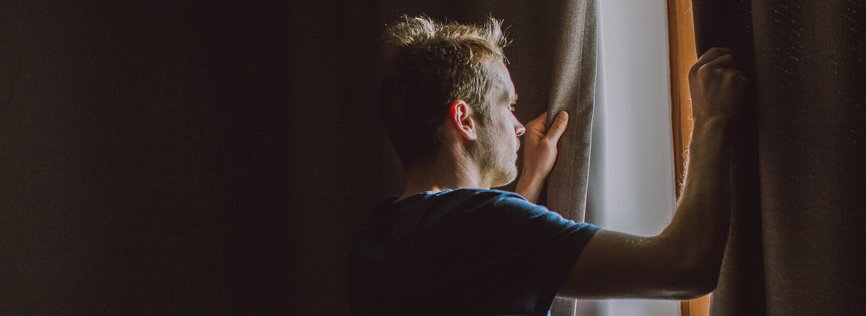 Das Bild zeigt einen Mann im kurzen Pyjama. Er steht an Fenster und ist im Begriff die Vorhänge zuzuziehen.