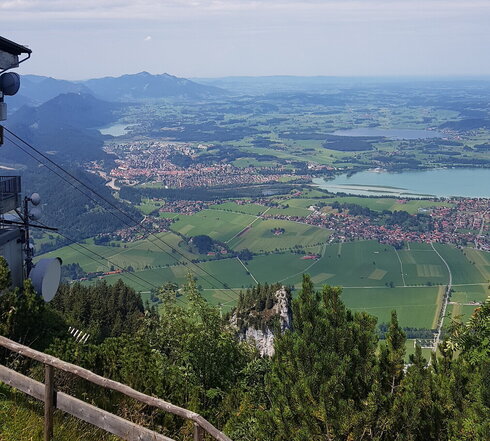 Das Bild zeigt die Aussicht von Tegelberg. Man sieht schier unendlich, bis zum Horizont über eine grüne, teilweise bewaldete Ebene. Vereinzelt sind unterschiedlich große Seen zu sehen. Der Himmel ist blau. Man sieht unzählige Gleitschirmflieger im Himmel. Die Sonne scheint. Im Vordergrund kann man einen Teil der Bergstation der Tegelbergbahn erkennen. 