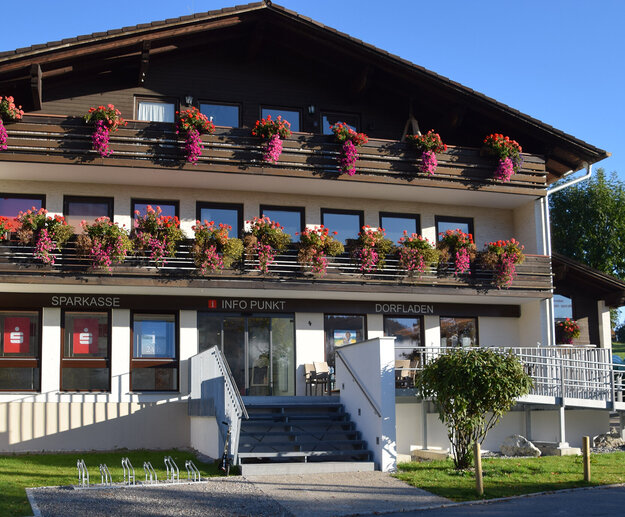 Das Bild zeigt ein zweistöckiges Gebäude mit zwei Balkonen, die jeweils an der Vorderseite des Gebäudes entlanglaufen. Die Balkone sind mit mehreren Blumenkästen bestückt, in denen üppig bunte Blumen wie Geranien und Petunien blühen. Eine mehrstufige Treppe führt zum Haupteingang. Über dem Haupteingang ist „Sparkasse, Info Punkt“ und „Dorfladen“ zu lesen. Das oberste Stockwerk des Hauses ist mit dunklem Holz verkleidet. Der Himmel ist blau. Die Sonne scheint.
