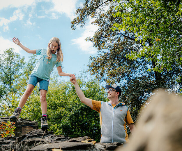 Das Bild fängt einen Moment der Freude und des Abenteuers zwischen einem Mann und einem jungen Mädchen ein. Sie stehen auf einer felsigen Erhebung, wobei das Mädchen spielerisch von der Kante springt. Der Mann, der eine gelbe Weste trägt, hält ihre Hand, um ihre Sicherheit zu gewährleisten. Das Mädchen, das ein blaues Hemd und Shorts trägt, schwebt in der Luft, ihre Arme ausgebreitet, als ob sie flöge. Der Hintergrund ist ein üppiger grüner Wald, mit Bäumen und Büschen, die das Landschaftsbild zieren. Der Himmel über ihnen ist ein klares Blau, das zur Gesamtstimmung des Bildes beiträgt. Das Bild strahlt Freiheit, Erkundung und die einfachen Freuden der Kindheit aus.
